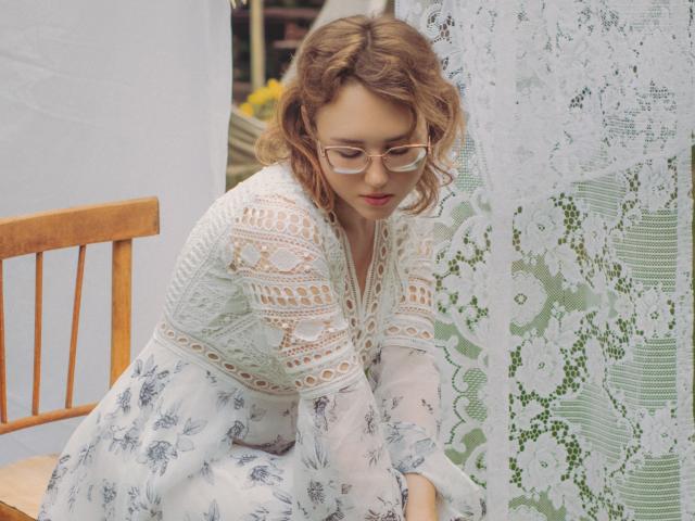 Teresa Birt, con cabello rubio rizado, gafas y una blusa blanca de encaje, apoyada en una silla de madera, falda blanca con estampado floral, cortina de encaje, mirando hacia abajo, luz suave, rostro bonito, linda..