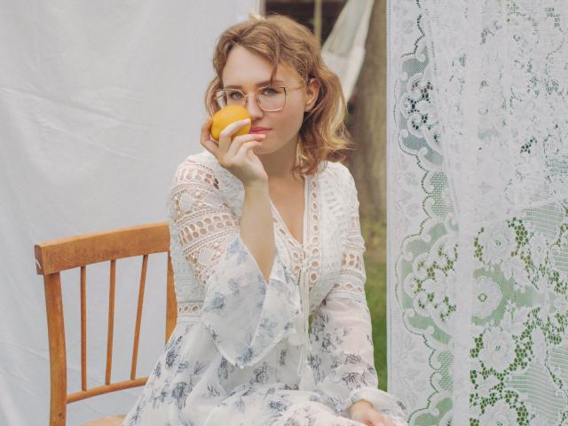 Teresa Birt, con cabello castaño ondulado, gafas y un vestido blanco de encaje, sosteniendo una manzana amarilla cerca de su boca, sentada en una silla de madera, con una cortina de encaje al fondo..