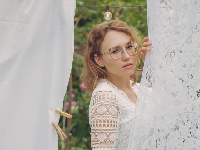 Teresa Birt, con cabello castaño ondulado, gafas y una blusa blanca de encaje, sosteniendo una cortina con un prendedor de madera, mirando por encima del hombro. Luz natural suave, sonrisa sutil..