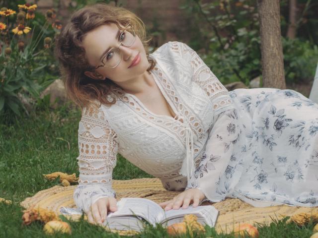 Teresa Birt, avec ses cheveux bruns bouclés, ses lunettes, son haut en dentelle blanche et sa jupe fleurie, est allongée sur une couverture jaune, en train de lire un livre. De l'herbe verte et des fleurs jaunes l'entourent..