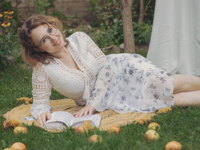 Teresa Birt, con gafas, cabello castaño ondulado, blusa blanca de encaje y falda floral, acostada sobre una manta de hierba, leyendo, rodeada de manzanas, sonriendo, atractiva, linda, luz suave..