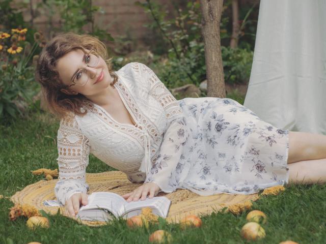 Teresa Birt, con el pelo castaño rizado y gafas, está acostada sobre una manta de hierba, vestida con una blusa blanca de encaje y una falda estampada con flores, leyendo un libro y con manzanas a su alrededor..