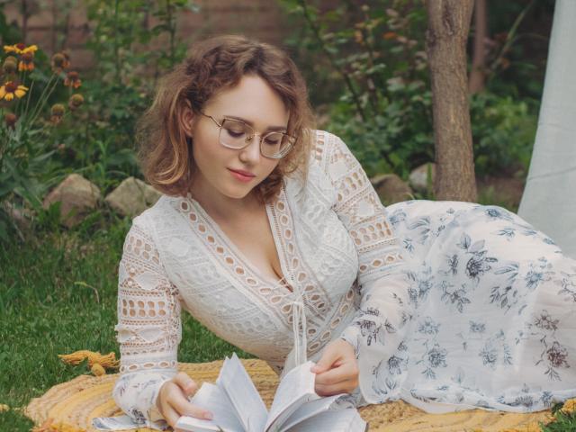 Teresa Birt, avec ses cheveux bruns bouclés, ses lunettes et sa robe blanche en dentelle, allongée sur l'herbe, en train de lire un livre, tenant des pages, avec une jupe blanche à motifs floraux, des fleurs jaunes, un arbre en bois, un jardin ensoleillé..