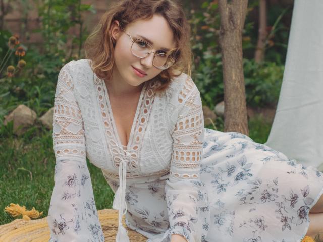 Teresa Birt, con cabello castaño rizado, gafas y un vestido blanco de encaje, apoyada en una cesta de mimbre, con sus senos visibles a través del vestido, en un jardín verde..