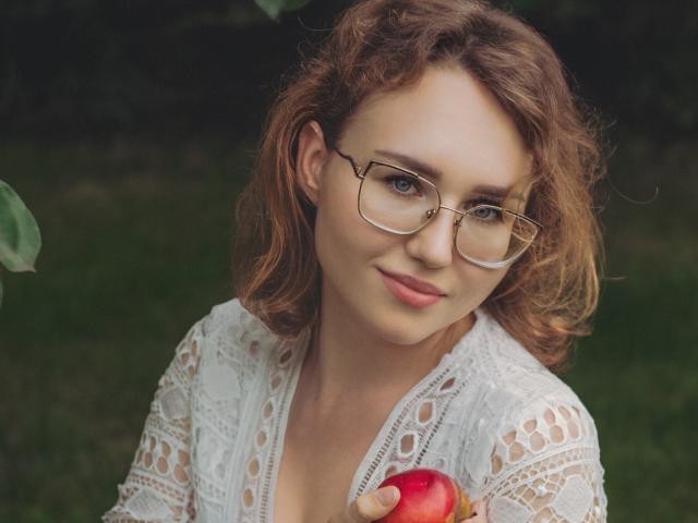Teresa Birt, con cabello castaño rizado, gafas, blusa blanca de encaje, sosteniendo una manzana roja, sonriendo suavemente, fondo de hierba, plano medio del torso, expresión amable, sosteniendo la manzana frente a ella..