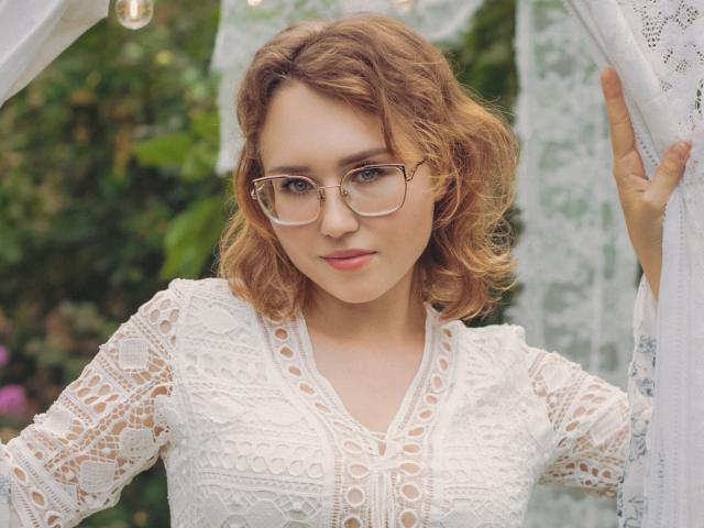 Teresa Birt, con cabello castaño rizado, usando gafas y un vestido blanco de encaje, sosteniendo una cortina, mostrando sus pechos a través del encaje. Sus glúteos no son visibles..
