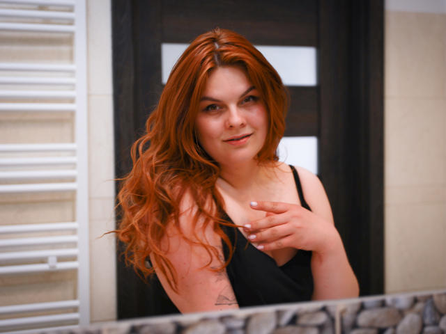 GingerKiss com cabelo ruivo, camiseta preta, tocando o peito, sorriso discreto, tatuagens no braço direito, suporte de toalhas branco à esquerda..