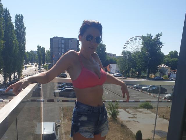 JoliDanielle ha la pelle abbronzata, piccoli seni in un reggiseno rosa e indossa pantaloncini di jeans. È all'aperto, con le braccia aperte e gli occhiali da sole, in un contesto urbano soleggiato..