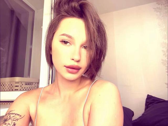 DarceyHeindl with short brown hair, light skin, and full pink lips, wearing a white spaghetti strap top, showing a tattoo on her right arm. Soft bedroom lighting.