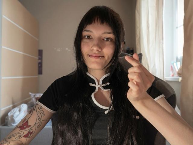 Valery Bazzle, con lunghi capelli neri, indossa una maglietta nera con bordi bianchi, tiene delle forbici nella mano destra, ha dei tatuaggi sul braccio sinistro, sorride alla telecamera, in una stanza illuminata dal sole..