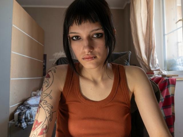 Valery Bazzle, con capelli neri, canottiera marrone, piercing al naso, piercing al labbro e tatuaggio sul braccio destro, che guarda verso la fotocamera, in una stanza illuminata dal sole..