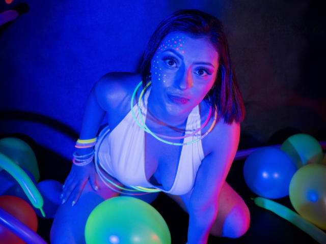 LizBonnet mit leuchtenden Punkten im Gesicht, trägt ein weißes Halstopp, bunte Armbänder und Neonlichter, kniet in einem dunkelblauen Raum mit bunten Ballons und zeigt ihre Brüste..