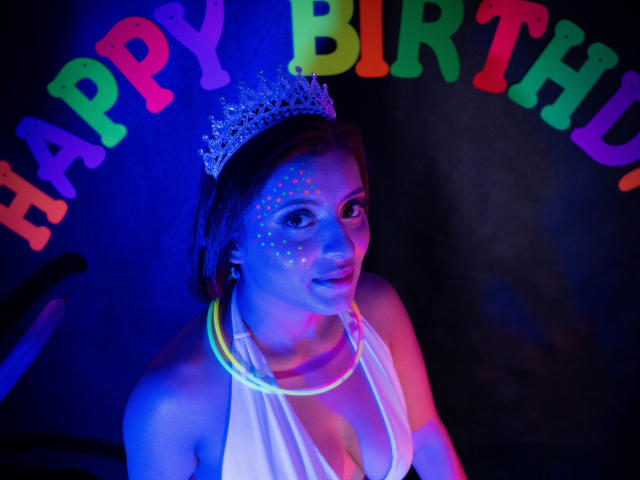 LizBonnet mit Neonlichtern im Gesicht, die eine glitzernde Krone und ein weißes Halstuch-Oberteil trägt, hat farbige Punkte auf ihrer Wange und eine leuchtende gelbe Halskette. Blaue und violette Beleuchtung..
