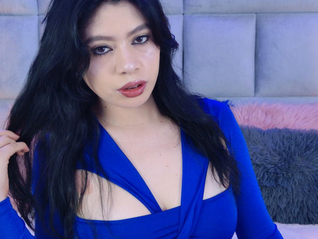 Linda Mancini, con cabello negro largo, vestida con un vestido azul con aberturas que muestran sus pechos, mirando a la cámara con lápiz labial rojo, con la mano en el cabello..