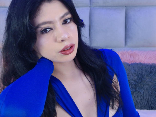 Linda Mancini, met lang zwart haar, draagt een blauwe jurk met een diepe halslijn, waardoor haar borsten zichtbaar zijn. Ze heeft roze lippen en een enigszins verleidelijke uitdrukking. Ze streelt haar haar..
