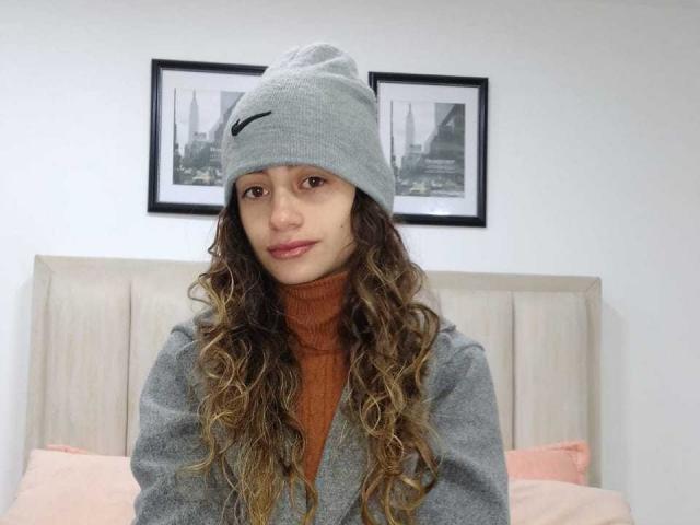Katalina Laurent mit langen, lockigen, braunen Haaren, die eine graue Mütze und einen braunen Rollkragenpullover sowie einen grauen Pullover trägt, sitzt auf einem Bett mit rosa Kissen und schaut in die Kamera..