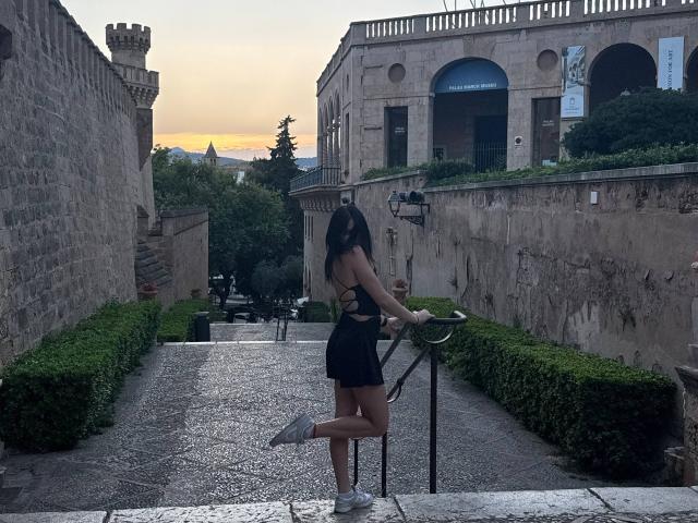 Nikksi, avec ses longs cheveux noirs, ses petites seins et sa jupe noire, se tient sur un chemin de pierre, tenant un vélo, portant des baskets blanches, et regardant en arrière un château au coucher du soleil..