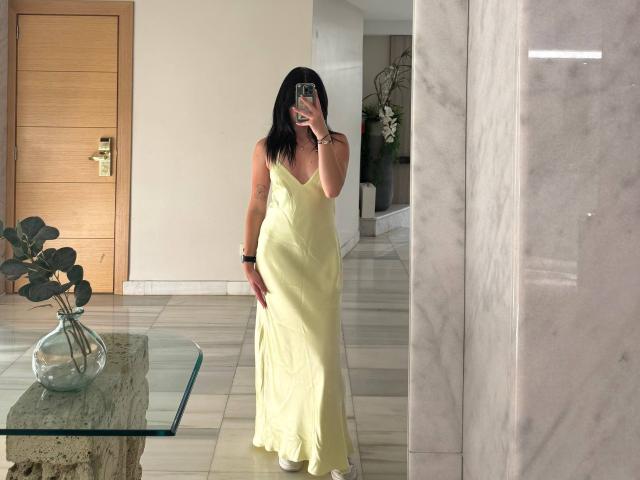 Nikksi, con i capelli neri, indossa un vestito giallo, tiene in mano un telefono, mostra il sedere, è in piedi di fronte a uno specchio, il riflesso mostra il seno, pavimento in marmo, tavolo in vetro..