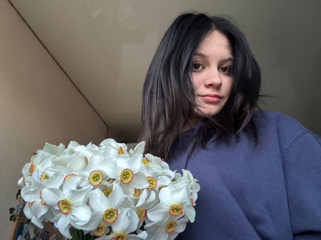 Nikksi, con i capelli neri, indossa una camicia blu e tiene in mano un bouquet di orchidee bianche con il centro giallo. Ha un'espressione seria, leggermente corrucciata..