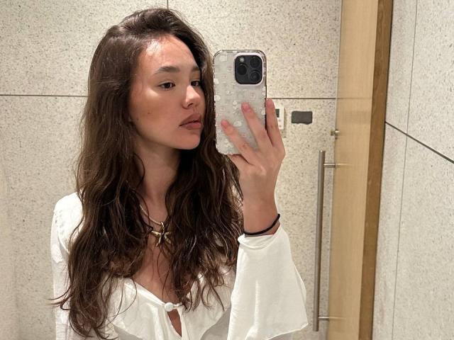 MyrtisTonti, avec de longs cheveux bruns ondulés, une blouse blanche, de petites seins, tenant un téléphone pour un selfie devant un miroir, un mur en carrelage beige, une porte en bois en arrière-plan, portant un bracelet noir, un maquillage discret..