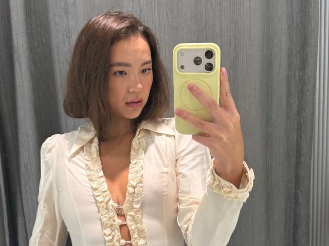 Myrtis Tonti mit kurzen, braunen Haaren, die ein weißes, gerafftes Hemd trägt und ein gelbes iPhone hält, macht ein Selfie im Spiegel und zeigt dabei ihre Brüste, in einem grauen Raum..