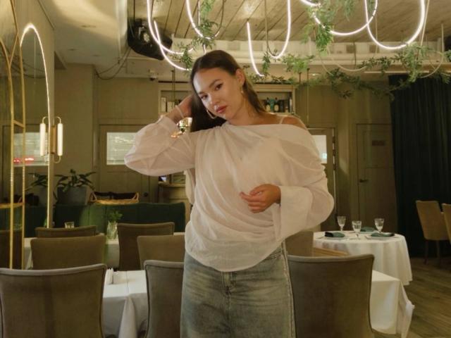 MyrtisTonti met lang bruin haar, een witte blouse met open schouders en blauwe jeans, staat in een restaurant, met haar hand in haar haar, terwijl ze een glas vasthoudt en naar de camera kijkt. Haar achterwerk is bedekt door de blouse..