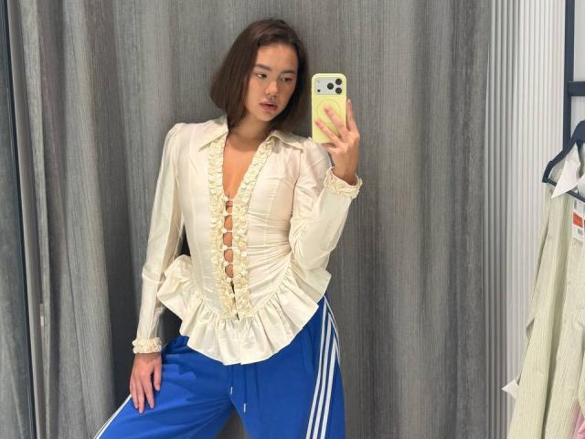 Myrtis Tonti, con i capelli castani corti, una camicetta bianca con volant e pantaloni da jogging blu, tiene in mano un telefono giallo e si trova davanti a delle tende grigie, guardandosi nello specchio..