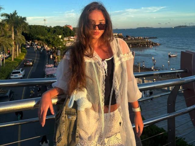 Myrtis Tonti, con lunghi capelli castani, che indossa occhiali da sole, una camicetta di pizzo bianco e una gonna di jeans, appoggiata a una ringhiera di metallo, tenendo una borsa bianca, sulla spiaggia al tramonto..