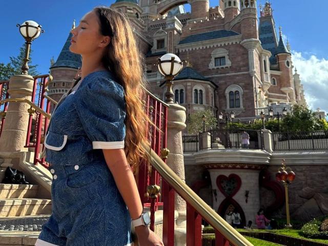 MyrtisTonti, com cabelos castanhos longos, vestindo um vestido azul com mangas bufantes, está em frente a uma escada, segurando um corrimão dourado, usando um relógio branco, em frente a um castelo..