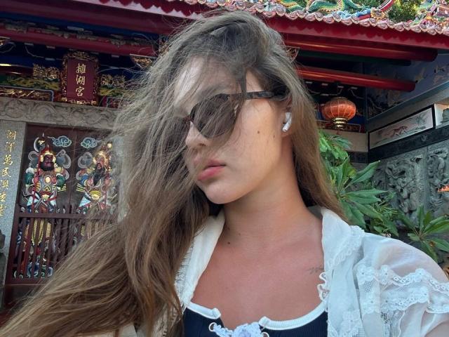 MyrtisTonti a de longs cheveux bruns qui flottent au vent, elle porte des lunettes de soleil sombres, une chemise blanche en dentelle et un haut noir, avec ses seins légèrement visibles, et elle a l'air sérieuse..