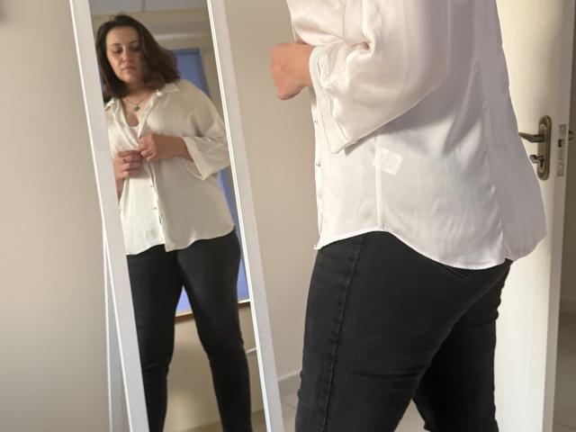 Elliance steht vor einem Spiegel, trägt ein weißes Hemd mit Knopfleiste und eine schwarze Hose und justiert ihr Hemd mit beiden Händen. Ihr Spiegelbild zeigt sie mit einer durchschnittlichen Figur..
