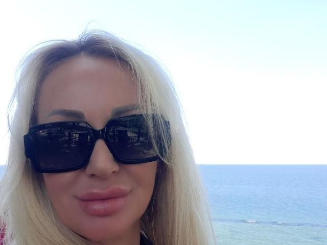 Ein geheimnisvolles Mädchen mit blonden Haaren, das schwarze Sonnenbrillen und rosafarbene Lippen trägt, lächelt in die Kamera und steht am Meer. Ihre Brüste sind nicht sichtbar..