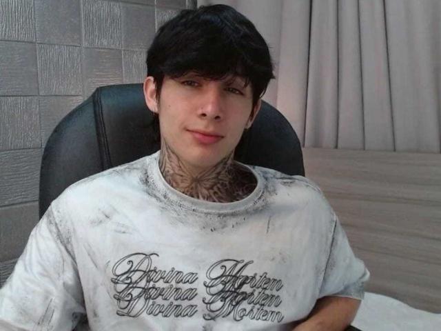 AkiraTattoo, con cabello negro corto, tatuajes en el cuello, vistiendo una camisa blanca sucia, sentada en una silla negra, sonriendo ligeramente..