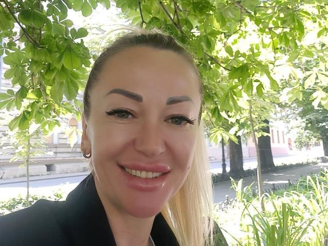 A MysteryLaddy tem cabelo loiro em um rabo de cavalo, sobrancelhas grossas, cílios longos, lábios rosados e brilhantes, e um casaco preto, sorrindo sob folhas verdes vibrantes..