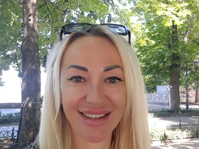 MysteryLaddy a les cheveux blonds, les yeux bleus, et elle sourit, avec un anneau de nez. Elle porte des lunettes de soleil noires sur la tête, et on voit un arbre et du soleil en arrière-plan..