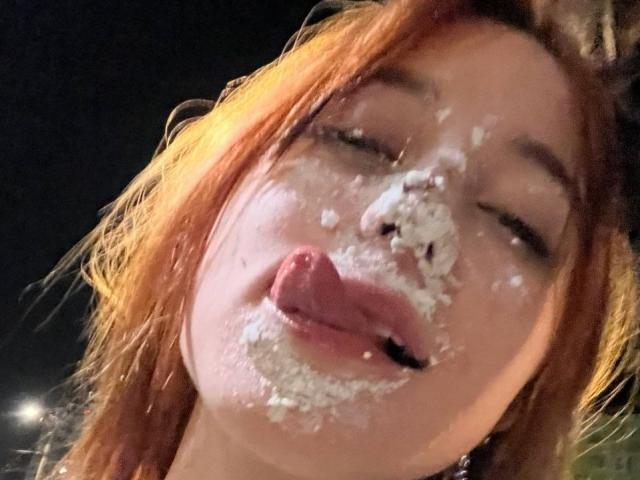 ArlenaStinnett with messy, white cream on her face, sticking out her tongue. She has orange hair and a slightly open mouth. Her eyes are half-closed. Nighttime.