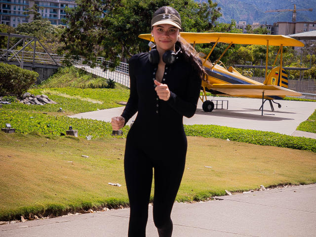 AlanaBlake, wearing a black bodysuit and visor, with headphones, stands in front of a yellow airplane, holding a finger up, smiling. Her ass is covered by the bodysuit.