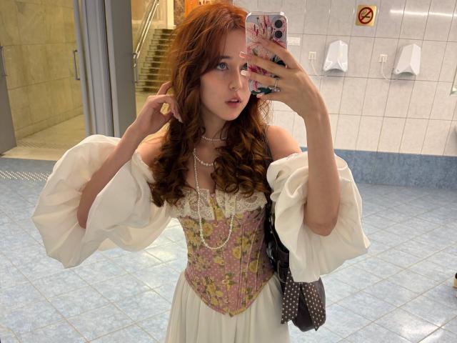 Arlena Stinnett, con i capelli rossi ondulati, indossa un abito bianco con spalline scoperte e maniche a sbuffo, un corpetto fiorito rosa e una collana di perle, e sta scattando un selfie in un bagno..