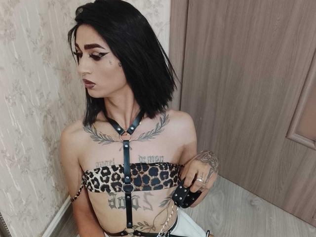IrisIrs with black hair, leopard-print strapless top, chest tattoos, and leather harness, holding a black polka-dotted leash, has dark makeup, and tattoos on her fingers.