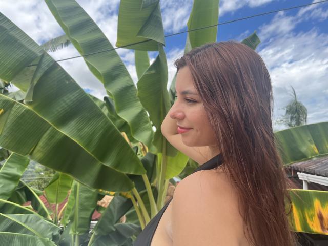 LinaFoxyHot, avec de longs cheveux bruns, de petites seins, et un haut noir à encolure américaine, regardant sur le côté avec un léger sourire, debout devant de grandes feuilles de bananier vertes..