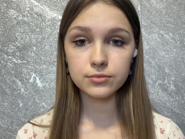 MelisaMagnie has long brown hair, light skin, green eyes, small nose, and is wearing a floral shirt with small pink flowers. She's looking straight at the camera with a neutral expression.