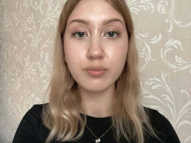LizetteAleyandrez has pale skin, green eyes, and blonde hair in a black shirt, wearing a small necklace with a white pendant. Her face is slightly shiny.