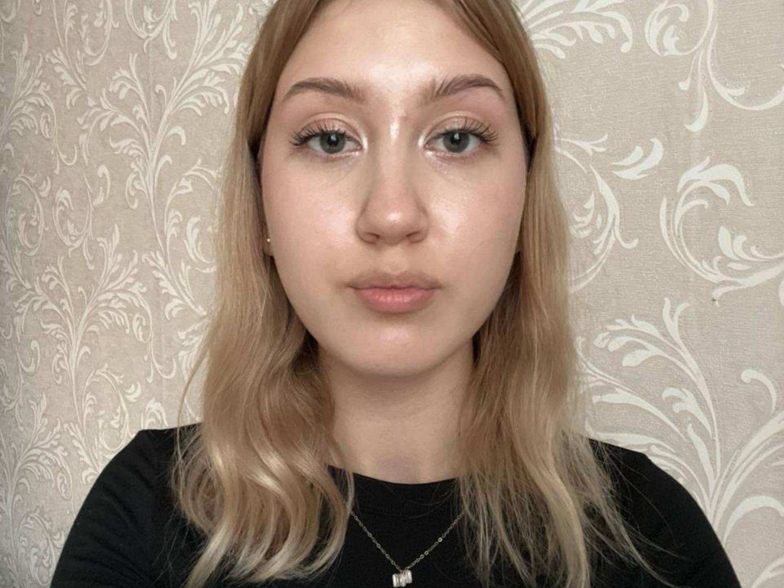 LizetteAleyandrez has pale skin, green eyes, and blonde hair in a black shirt, wearing a small necklace with a white pendant. Her face is slightly shiny.