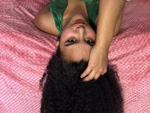 AriaVega, com cabelos pretos encaracolados, está deitada em uma cama rosa com textura, vestindo uma blusa verde, com a cabeça apoiada na mão, olhando para cima com batom vermelho e maquiagem discreta..