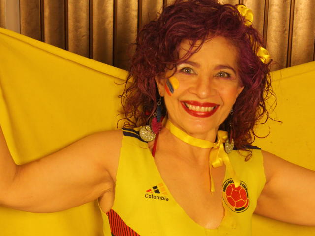 Dalila Najjar met krullend rood haar, een gele sporttop, gele schmink op haar gezicht, rode lippenstift, lachend, een gele lintketting, een gele vlag, die ze omhoog houdt, met kleurrijke oorbellen, en een blijk van geluk..