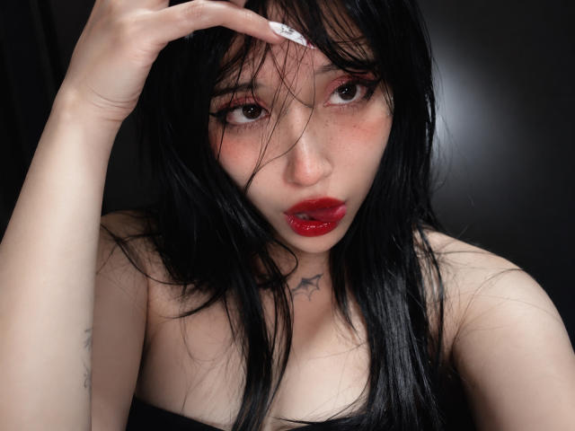 Violett: ragazza con capelli neri, labbra rosse e lentiggini, che guarda la fotocamera, con una mano tra i capelli, un tatuaggio a forma di stella sul petto, indossa una maglietta nera, pelle chiara, seno piccolo..