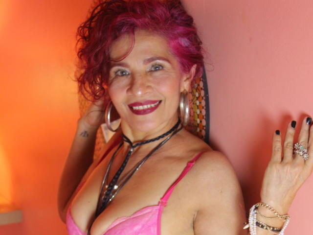 Dalila Najjar mit rosa Haaren, Reifenohrringen, schwarzem Nagellack, einem rosa BH, lächelnd, die eine schwarze Halskette und Ringe präsentiert, hält eine Hand mit schwarzen Nägeln hoch..