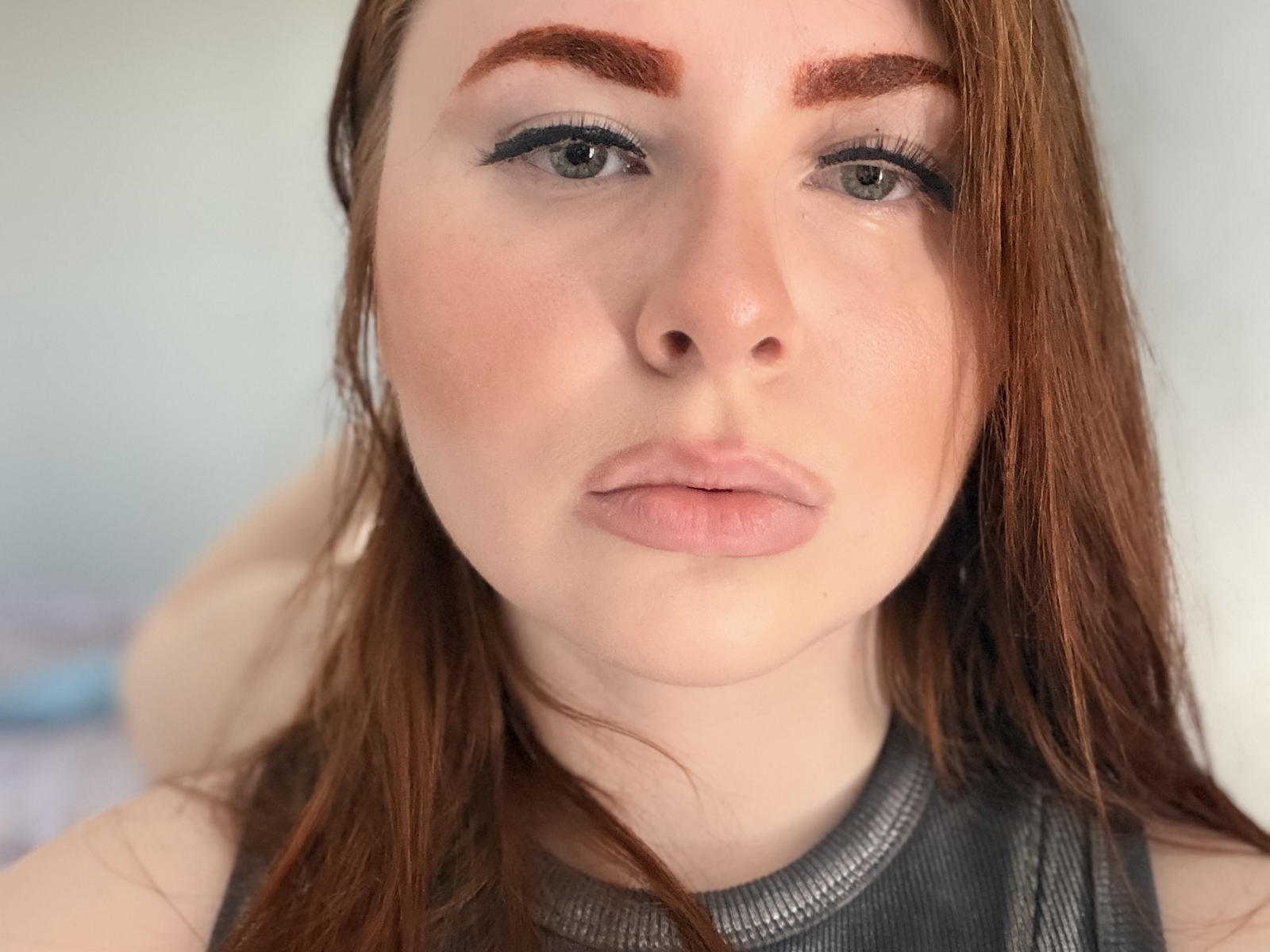 ShenitaSingh has pale skin, green eyes, brown eyebrows, long red hair, and is wearing dark eyeliner and pink lipstick. Her lips are slightly parted. She's wearing a gray top.