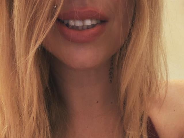 Il viso di XimenaBella, con le sue labbra rosse e i denti bianchi, i capelli biondi che le coprono gli occhi, un piccolo piercing al naso e un solo orecchino sul lato sinistro..