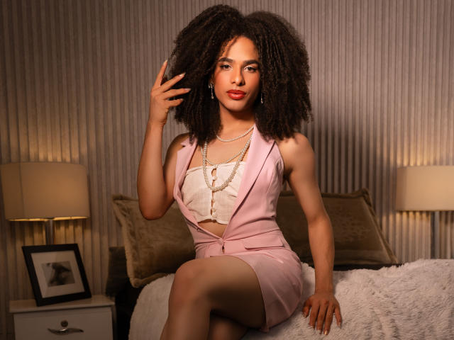 Kataleya Fowler mit einer großen, lockigen Frisur, einem pinken Anzug, einem weißen Oberteil, einer Perlenkette und rotem Lippenstift, sitzt auf dem Bett, eine Hand im Haar, die Beine gekreuzt, wirkt selbstbewusst und attraktiv..
